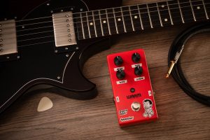 Warm Audio Dirty Boy Jr. Booster pedal next to a guitar on a table.