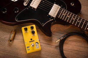 Warm Audio Dirty Boy Jr. Fuzz pedal next to a guitar on a table.